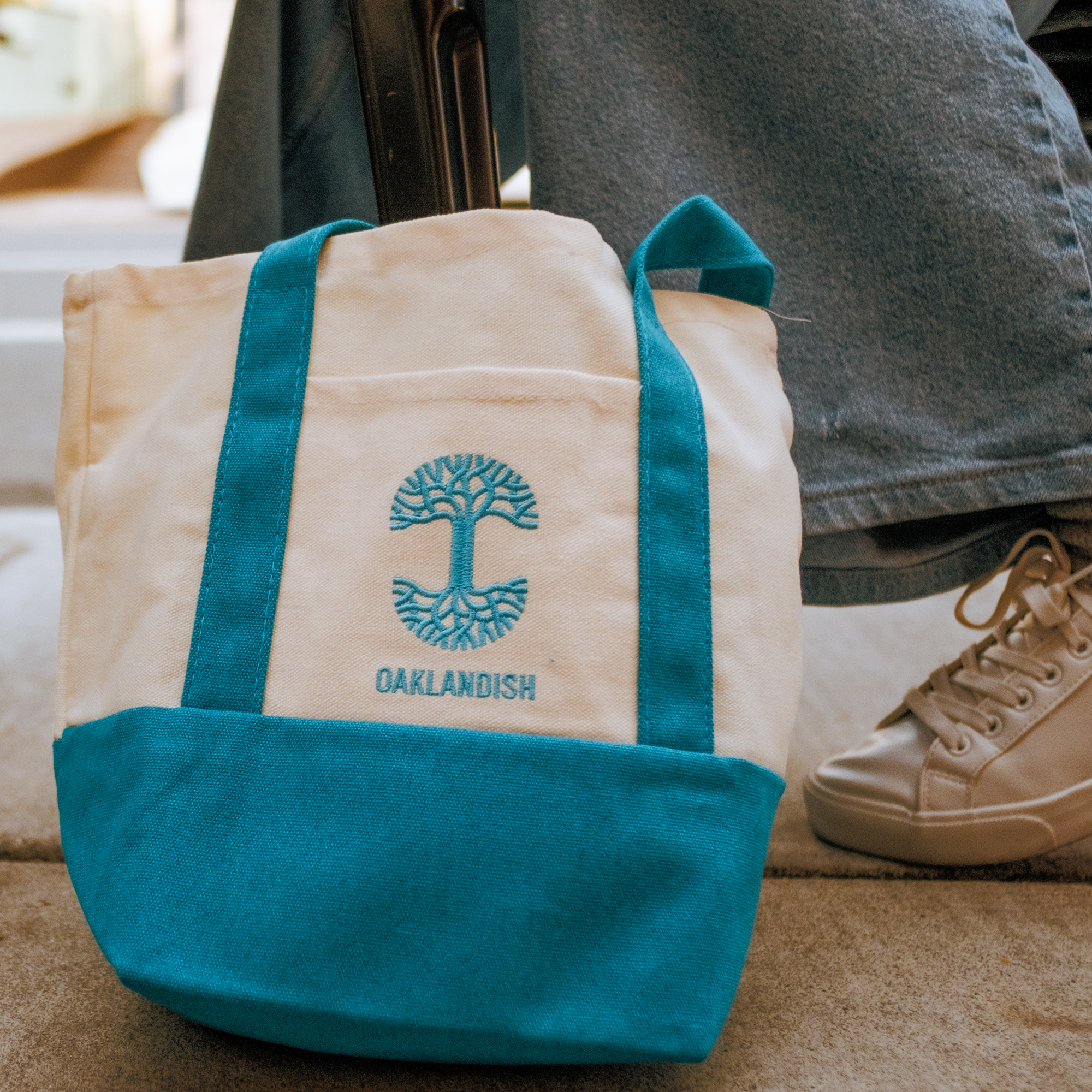 The Oaklandish Mini Grocery Tote, a white canvas bag with bright blue handles and base, features an embroidered tree logo and “OAKLANDISH” in blue. A person in denim jeans and beige sneakers stands beside the Oaklandish accessory.