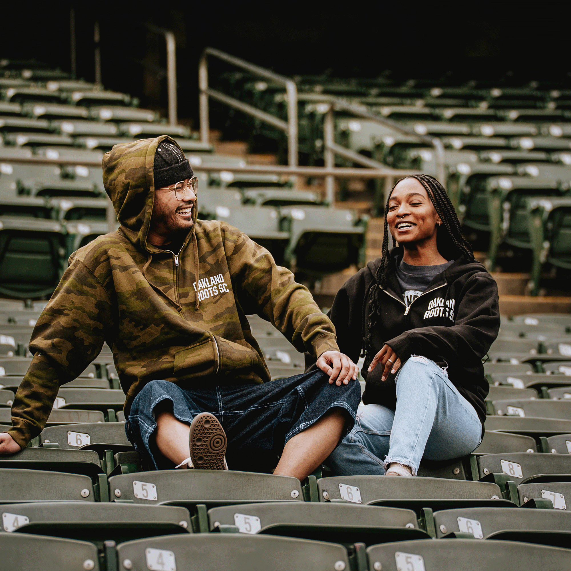 A smiling man in an Oakland Roots SC Classic Zip Hoodie and a woman in a black hoodie lounge on green stadium seats, laughing together. Both sport casual outfits and sneakers amid empty seats at the lively Oakland Roots SC venue.