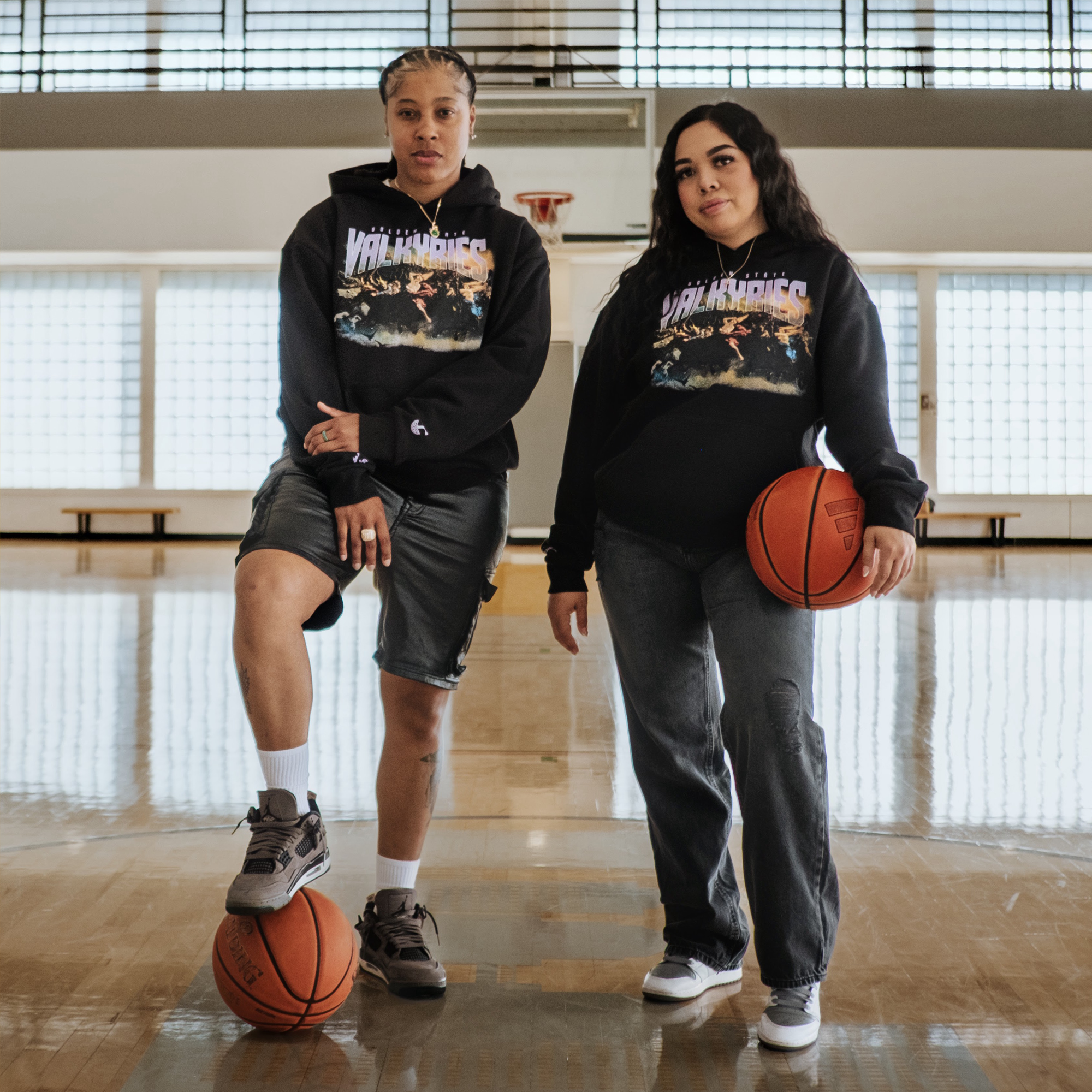 Two women stand confidently in a sunlit gym, each wearing black GSV Odin Hoodies. One, in shorts, rests a foot on a basketball; the other wears jeans and holds a ball.