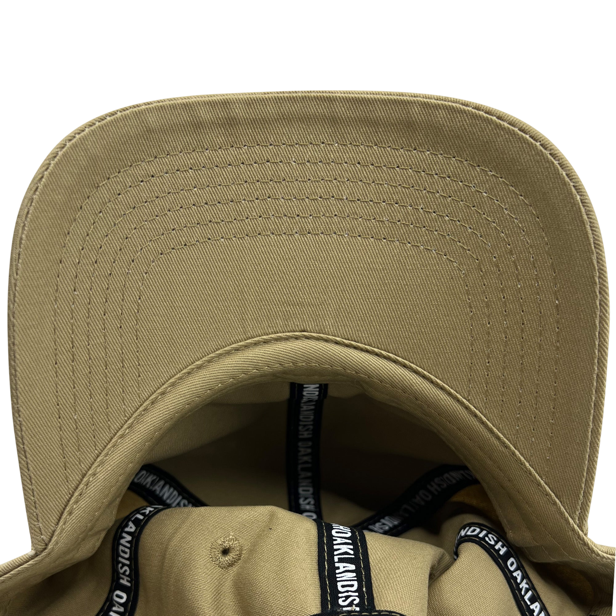 A close-up view of the inside of the Official Micro Logo Dad Hat by Oaklandish, showcasing its beige color, curved brim, and precise stitching. The inner lining is adorned with black bands featuring white text that repeatedly spells "Oaklandish" around the edges. The sturdy fabric and clear stitching are evident throughout, along with an adjustable strap for a perfect fit.
