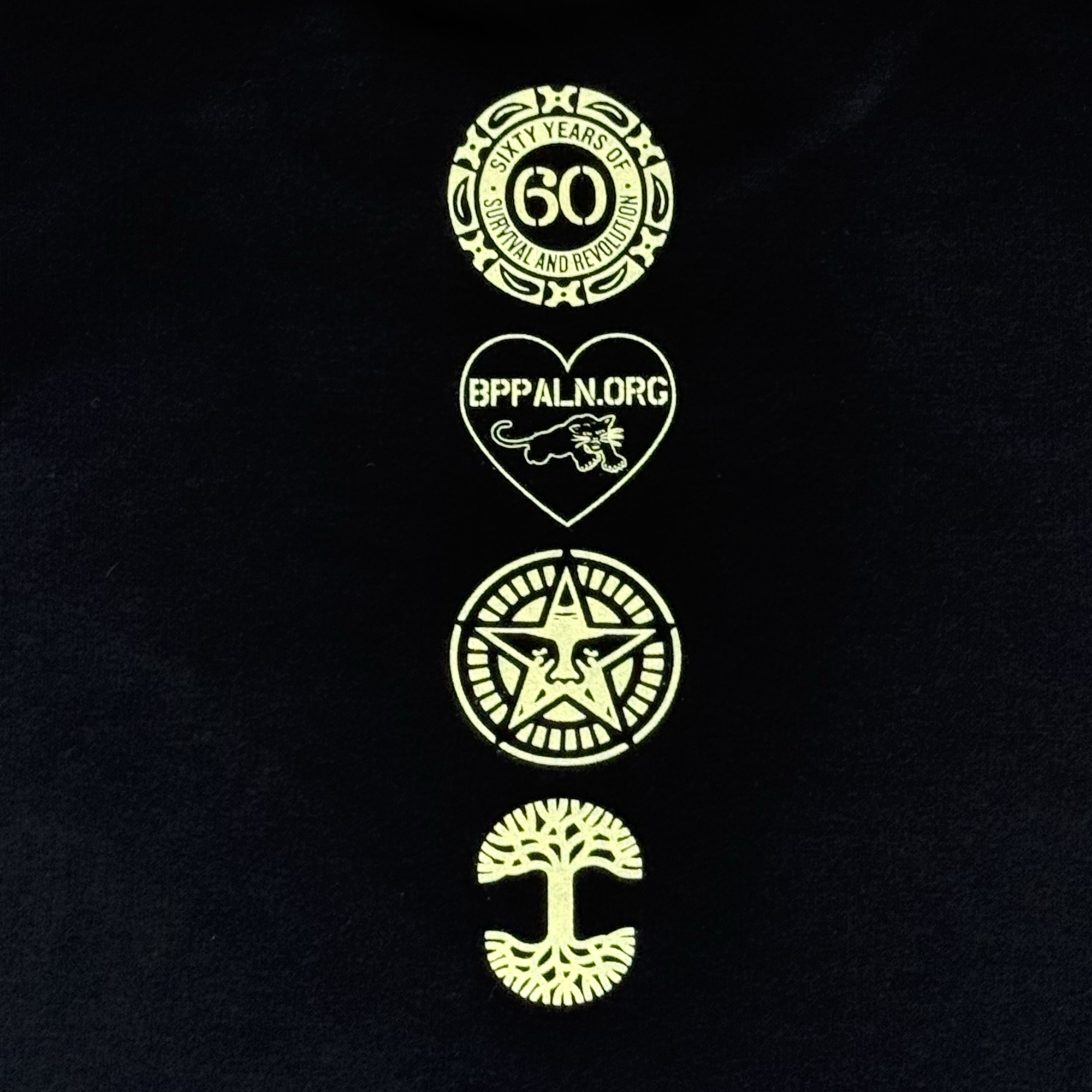 The Obey x BPPALN x Oaklandish 60th Educate to Liberate Tee from Oaklandish features a black background with four yellow logos: a 60th anniversary emblem, heart with panther and "BPPALN.ORG," star with eagle, and a tree with roots and branches.