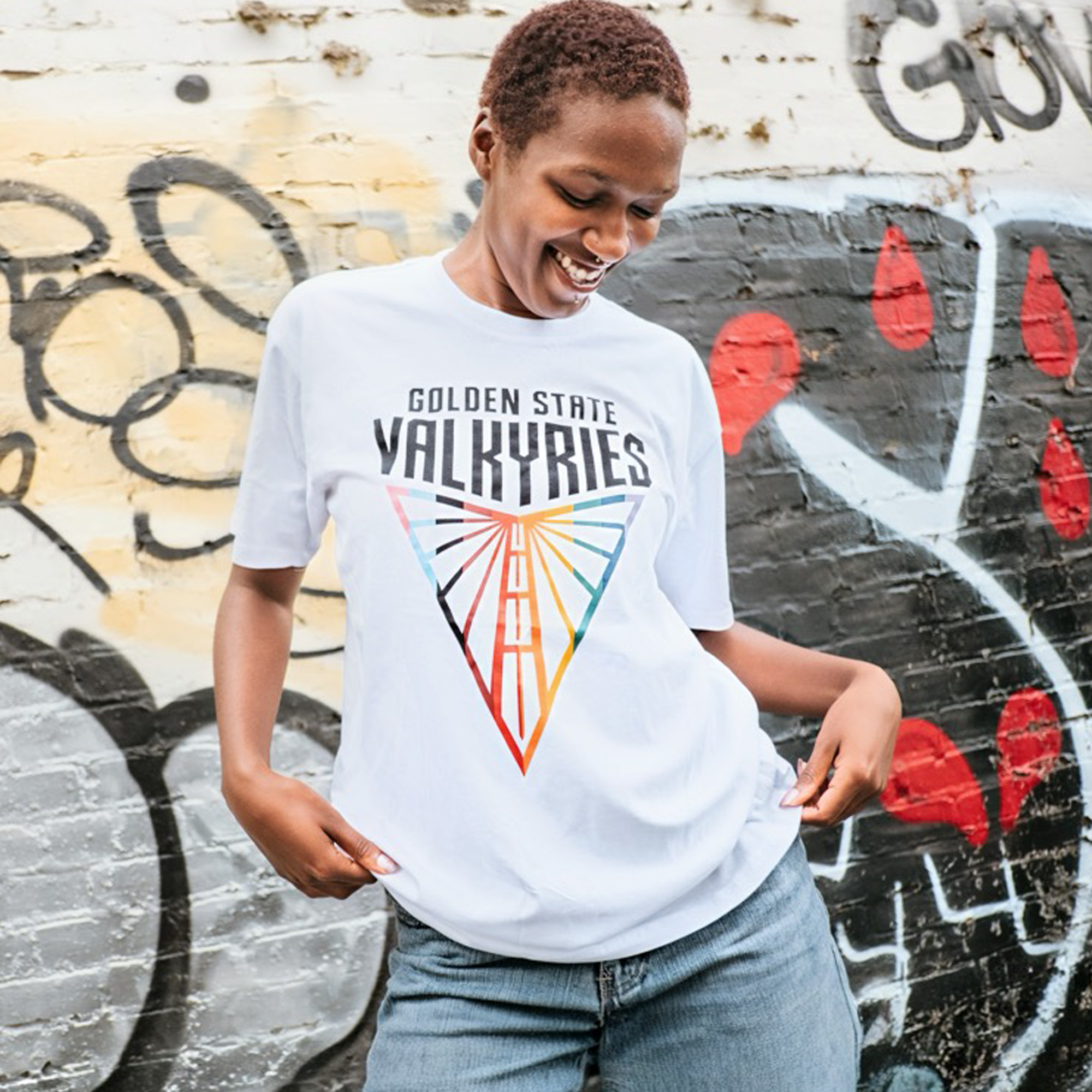 A smiling person with short hair wears the GSV Logo Love Tee by GSV, featuring a vibrant "Golden State Valkyries" geometric design. Made from 100% cotton, they stand before graffiti, holding the hem and looking down in a joyful pose.