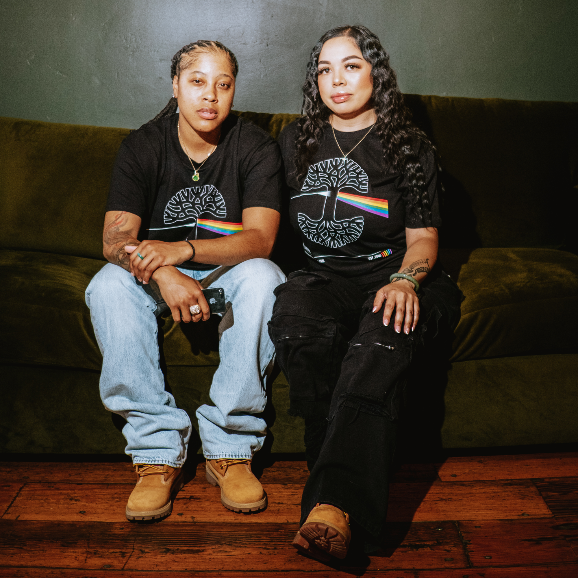 Two people sit closely on a dark green couch in Oakland, both wearing Oaklandish Prismatic Tees—100% cotton classic fit shirts featuring a white tree and rainbow design. One pairs it with light jeans and tan boots, the other with black pants and shoes.