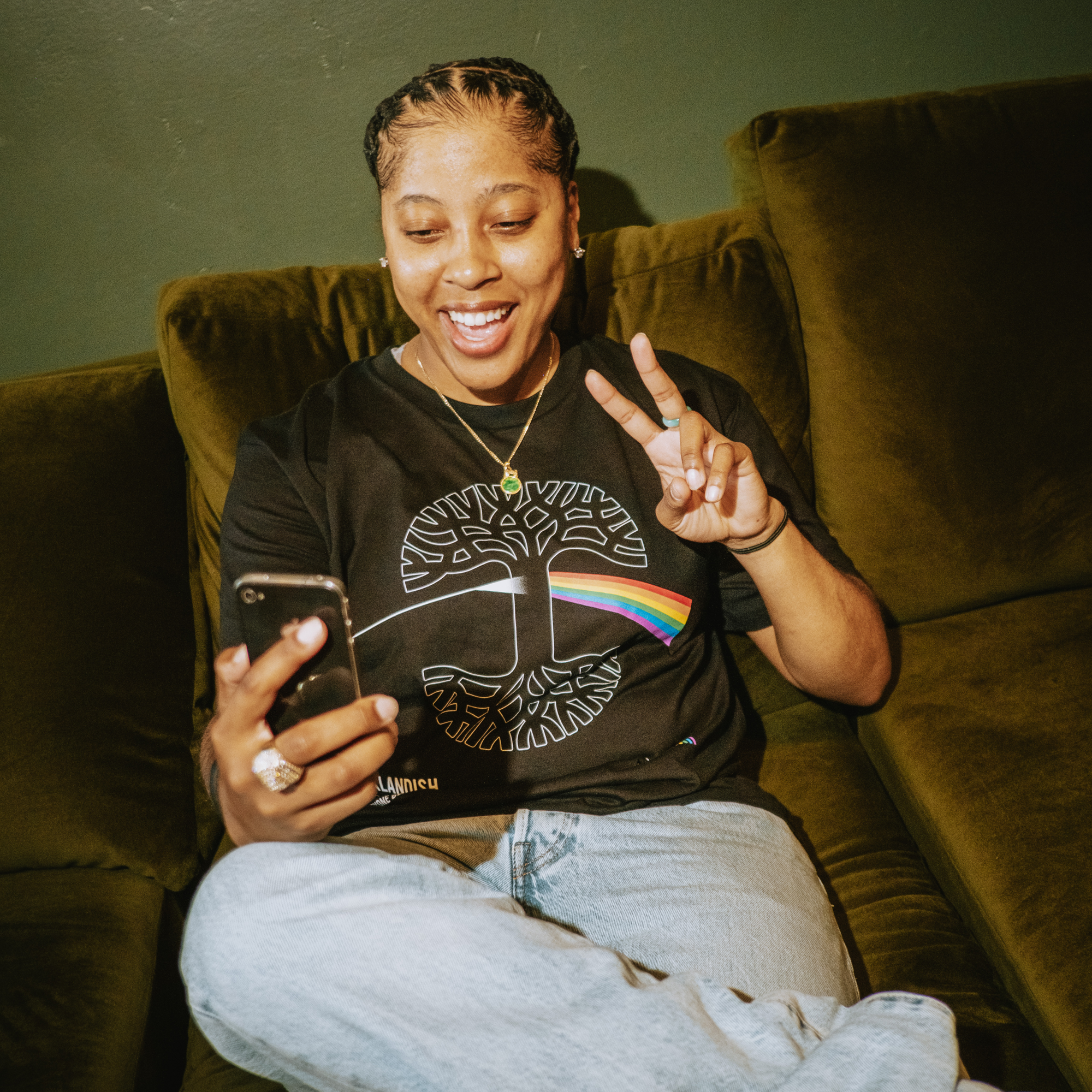 In Oakland, someone smiles and flashes a peace sign while checking their phone, seated on a green velvet couch. They wear the Oaklandish Prismatic Tee—100% cotton with a rainbow and tree design—paired with light jeans against a dark green wall.