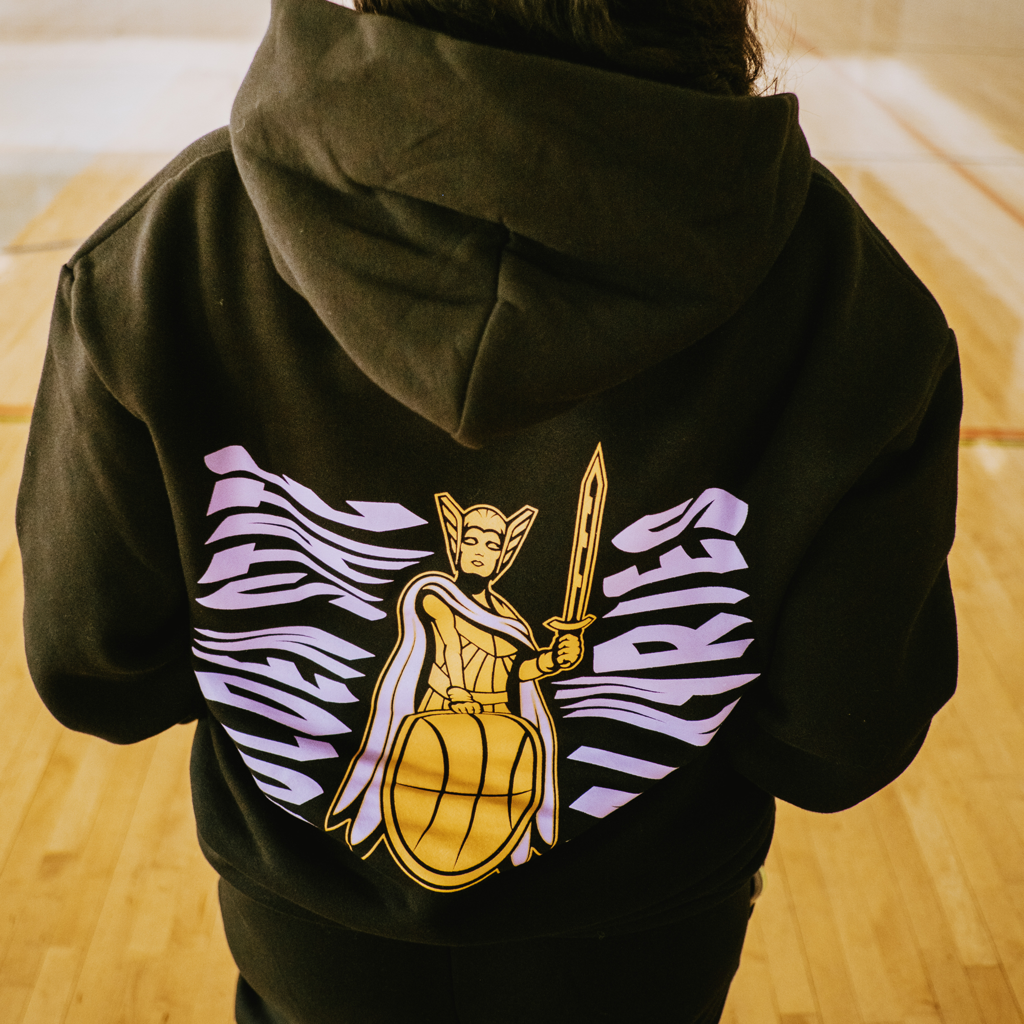 In a gym, a person wears the GSV Renowned Hoodie by GSV, featuring a warrior with sword and shield flanked by purple wings under "UNREAL ATHLETICS"—inspired by the Golden State Valkyries and available in men's sizing.