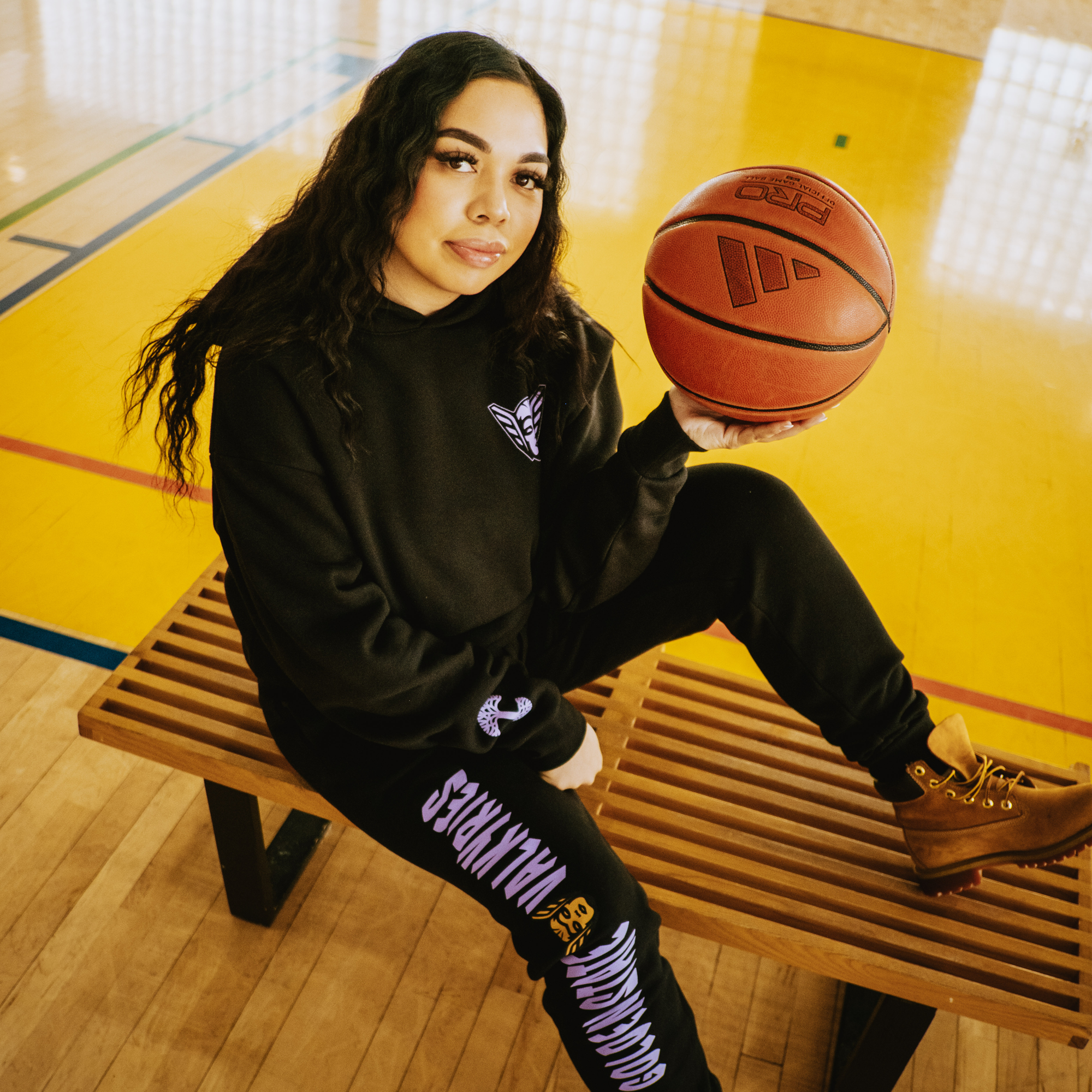 A woman with long wavy hair sits on a gym bench, holding a basketball. She wears GSV Renowned Pant and a Valkyries hoodie with white lettering. The yellow basketball court floor is lined for play.