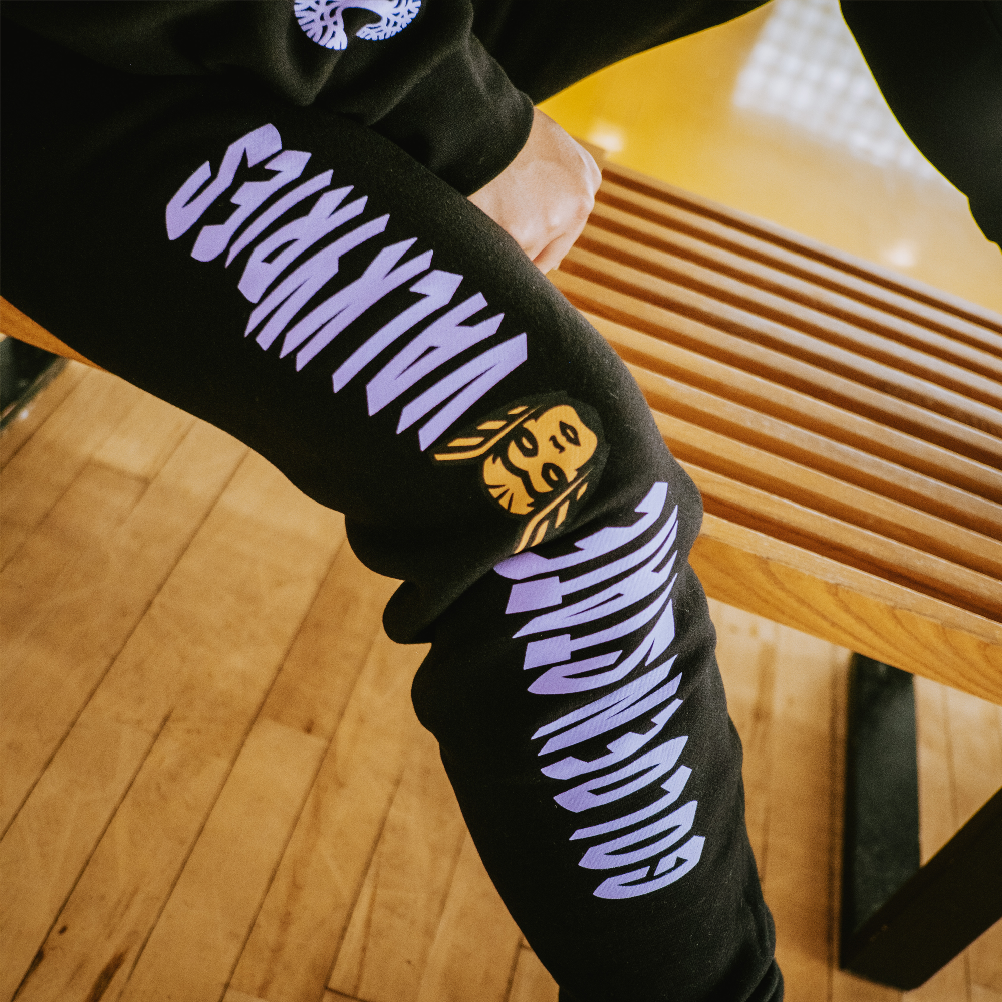 A person wears GSV Renowned Pant by GSV in men's sizing, with bold lavender "GOOGLY EYED MALWARE" text and a yellow cartoon face, sitting on a wooden bench on a gym floor—only their lower body and hand visible.