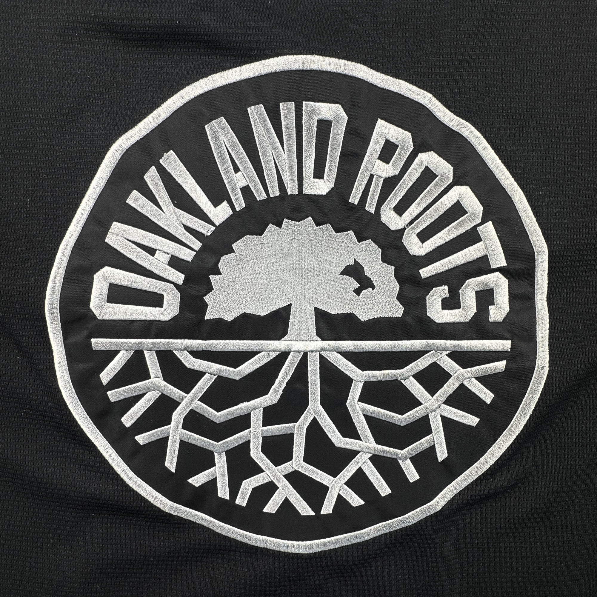 Circular black patch with "OAKLAND ROOTS" in bold white letters. At the center is a white tree with spreading roots and a small bird flying near the branches. Inspired by the Oakland Roots SC, this bold, graphic emblem is perfect for showing local pride on the ORSC Button Up Jersey by Oakland Roots SC.