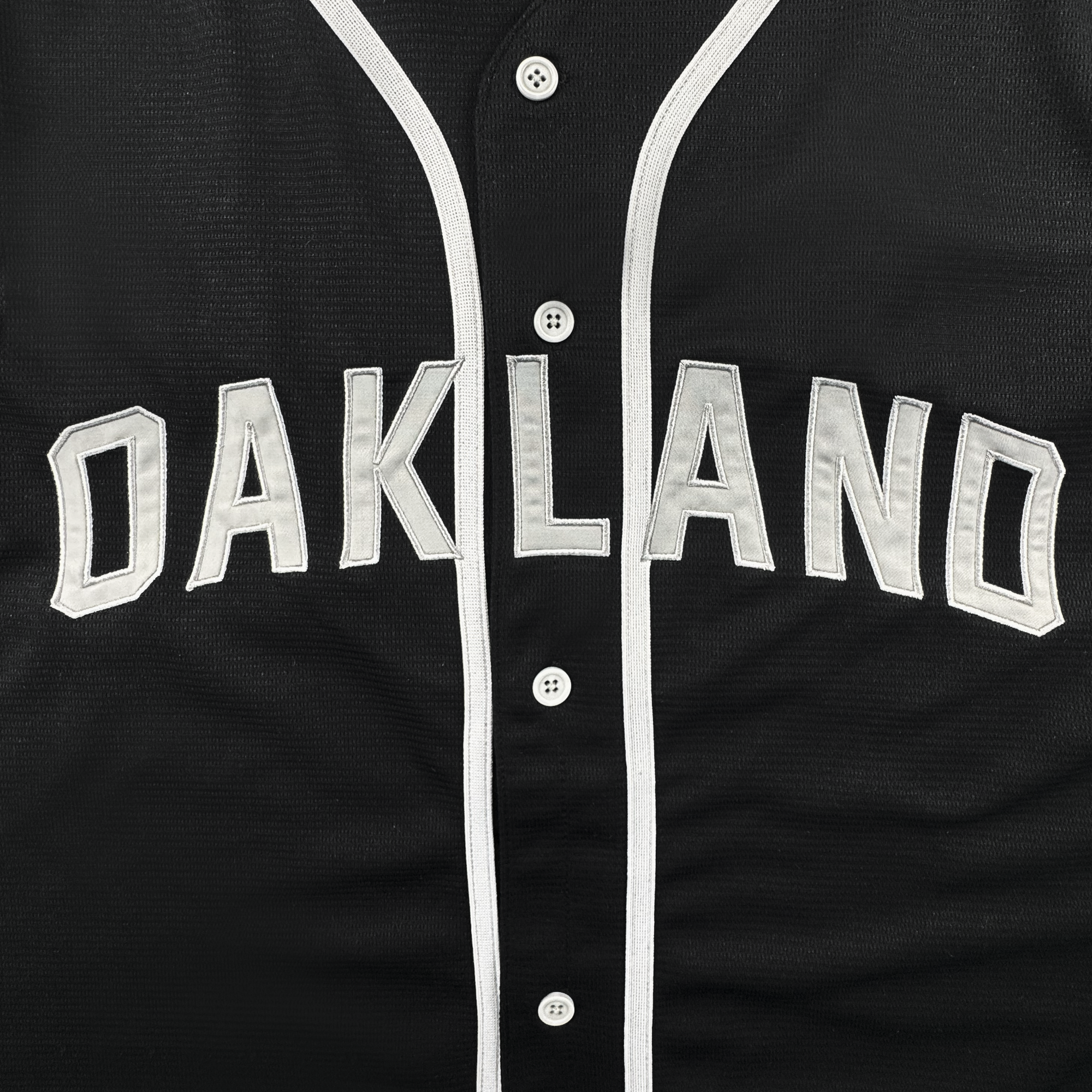A close-up of the ORSC Button Up Jersey by Oakland Roots SC, featuring bold white "OAKLAND" lettering outlined in silver across the chest. This black jersey showcases the signature Oaklandish Lifestyle style with white buttons down the front and white trim along the button panel.