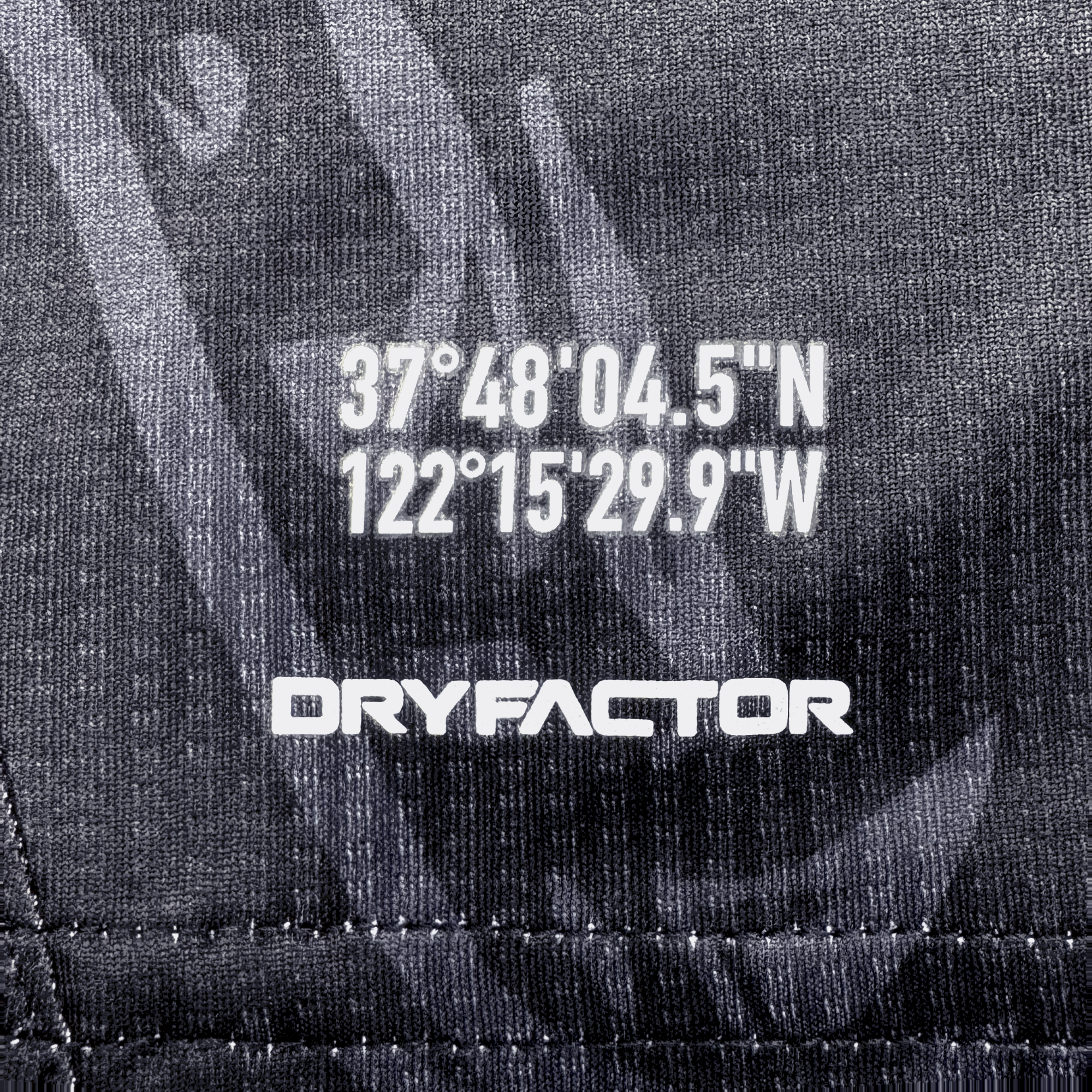 Close-up of dark textured fabric from the Unisex Oak-ness Awakens Kit by Oakland Roots SC. White coordinates "37°48'04.5"N 122°15'29.9"W" appear above bold "DRYFACTOR," with stitched lines and patterns inspired by Lake Merritt and Oakland folklore.