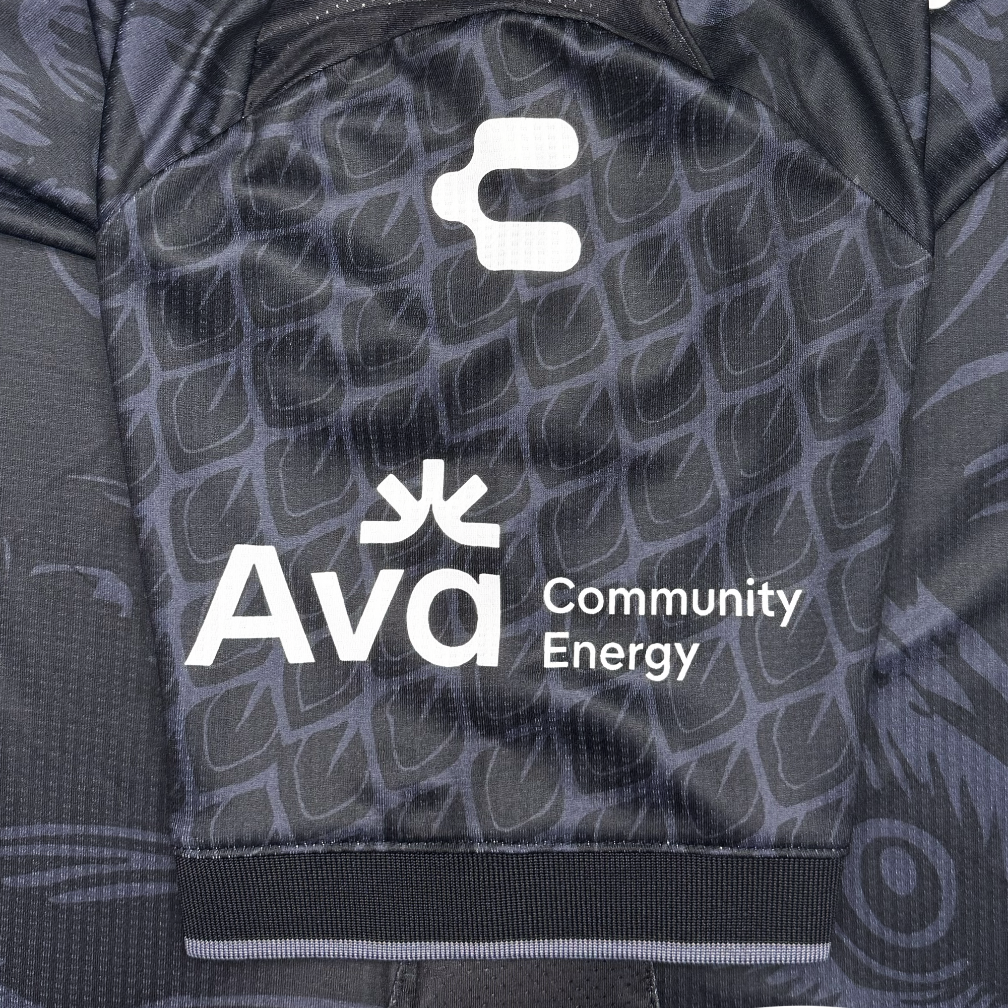 A close-up of the Oakland Roots SC Custom Unisex Oak-ness Awakens Kit, a black jersey with a subtle geometric pattern. The sleeve features white “C” and “Ava Community Energy” logos, with Ava’s sunburst celebrating Oakland folklore and Lake Merritt.