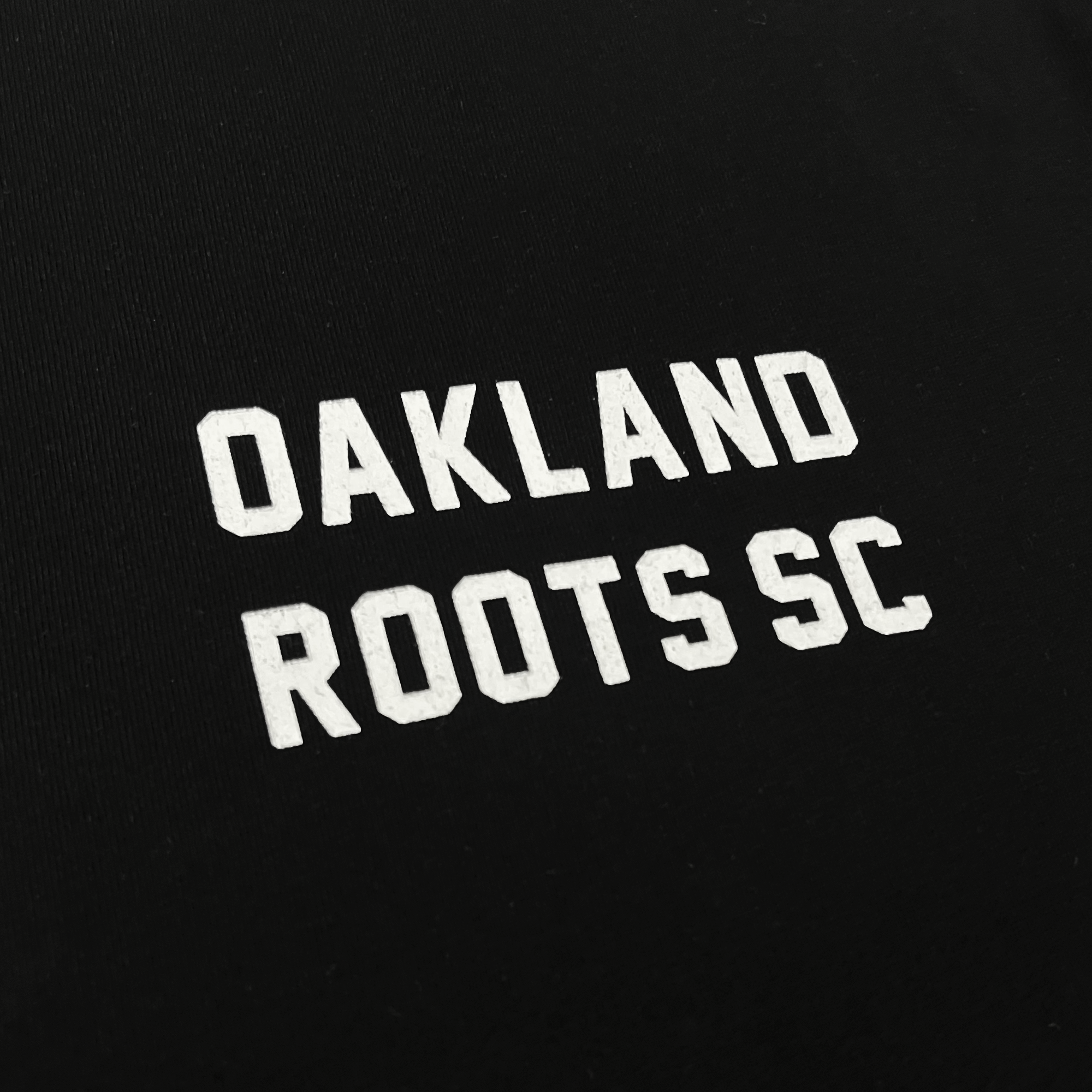 A close-up of the Oakland Roots SC Classic 2.0 LS Tee, a black long sleeve t-shirt featuring the words "OAKLAND ROOTS SC" printed in bold, uppercase white letters. The text is centered and stands out starkly against the dark background, embodying the spirit of this dynamic sports club.
