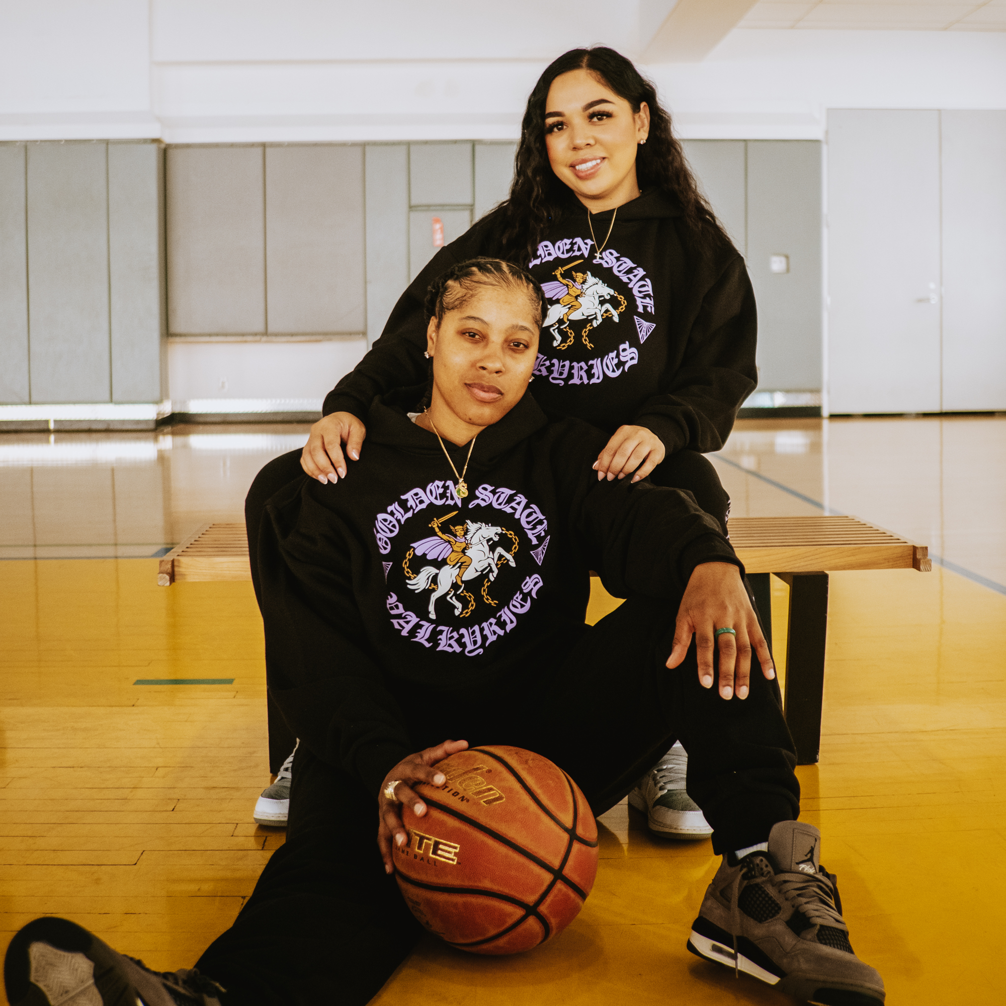 Two women pose in a gym with wooden floors and gray walls, both wearing GSV ROTV Hoodies by GSV (men’s sizing) and sneakers. One sits on a bench, the other on the floor holding a basketball.
