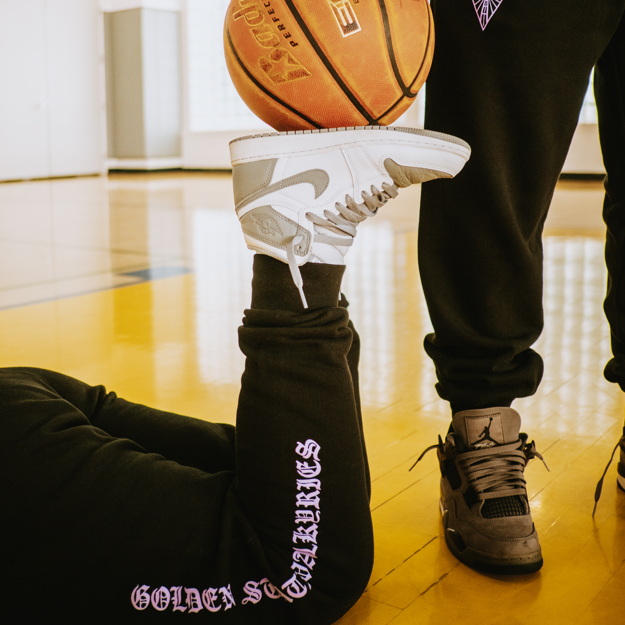 On a basketball court, someone in GSV ROTV Pants balances a white Nike Air Jordan 1 on their foot while lying down, as another person stands nearby in matching GSV sweatpants and black sneakers with a basketball floating above.