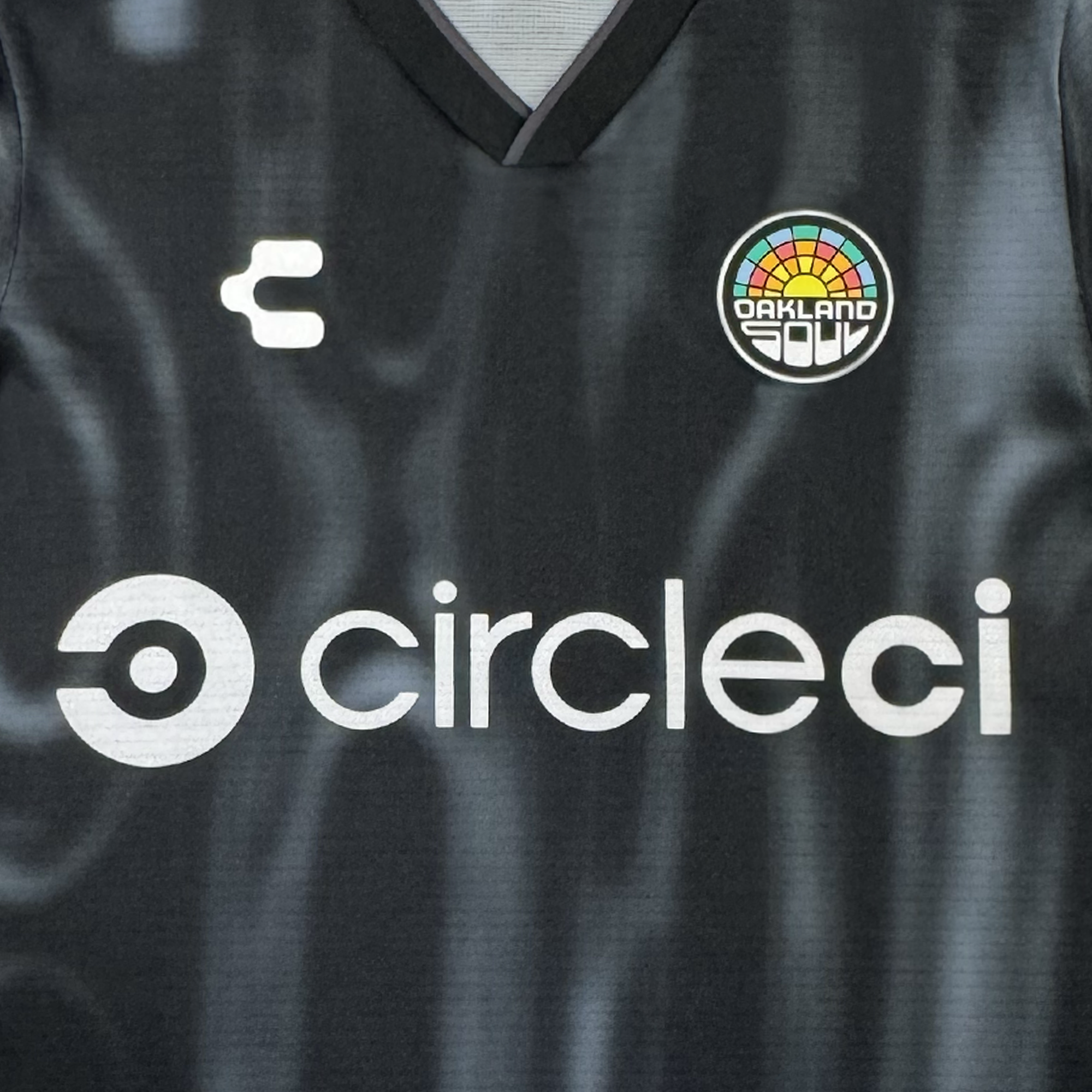 The Custom 2026 Oakland Soul SC Spirit Jersey by Oakland Soul is black with a white “circles” logo and text at the center, a stylized “C” on the left chest, and the colorful circular Oakland Soul crest on the right. The fabric features a subtle wavy texture.