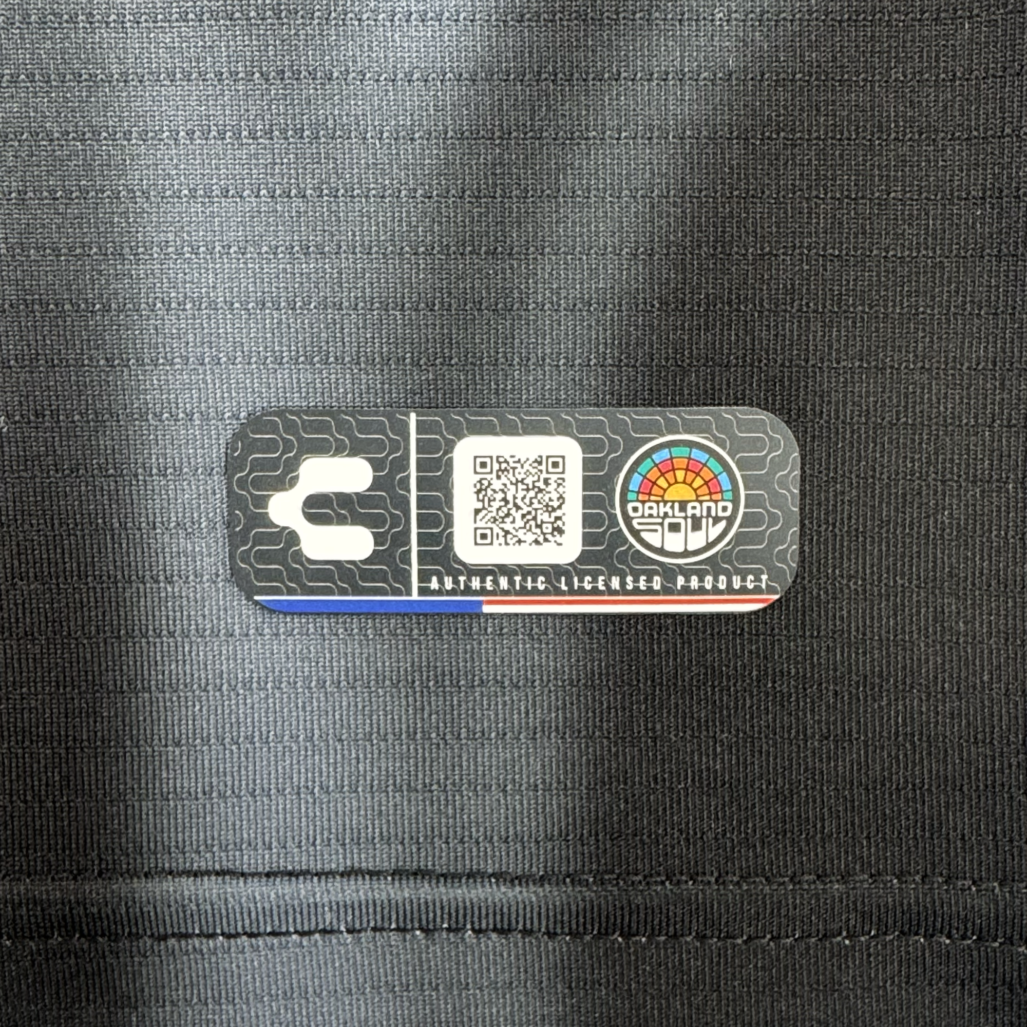 A close-up of the Custom 2026 OAKLAND SOUL SC SPIRIT JERSEY tag shows a QR code, stylized “C,” rainbow Oakland Soul logo, and “AUTHENTIC LICENSED PRODUCT” on silver and black with visible thin fabric lines.