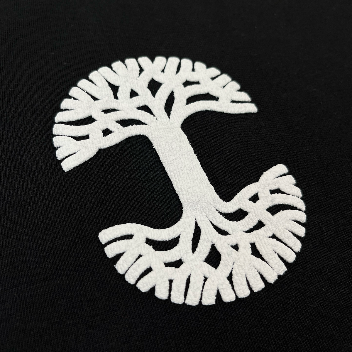 The Temple LS Tee by Oaklandish features a white embroidered, stylized tree design on black fabric, with branches and roots forming a kidney-like circular shape. Crafted from 100% heavy-weight cotton for lasting comfort and durability, this unique artwork is showcased on a men’s long sleeve t-shirt.
