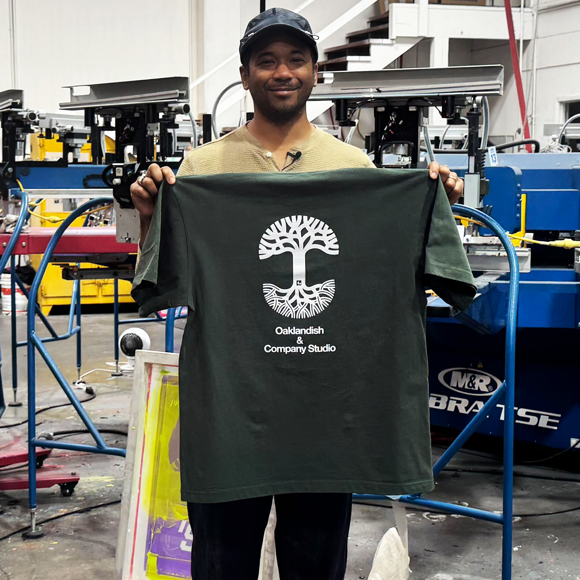 In a print shop, someone holds The East Bay Tee by Oaklandish—a dark green, limited-edition shirt featuring a white tree graphic and the words “Oaklandish & Company Studio,” honoring East Bay pride. Large printing machines and colorful materials fill the background.