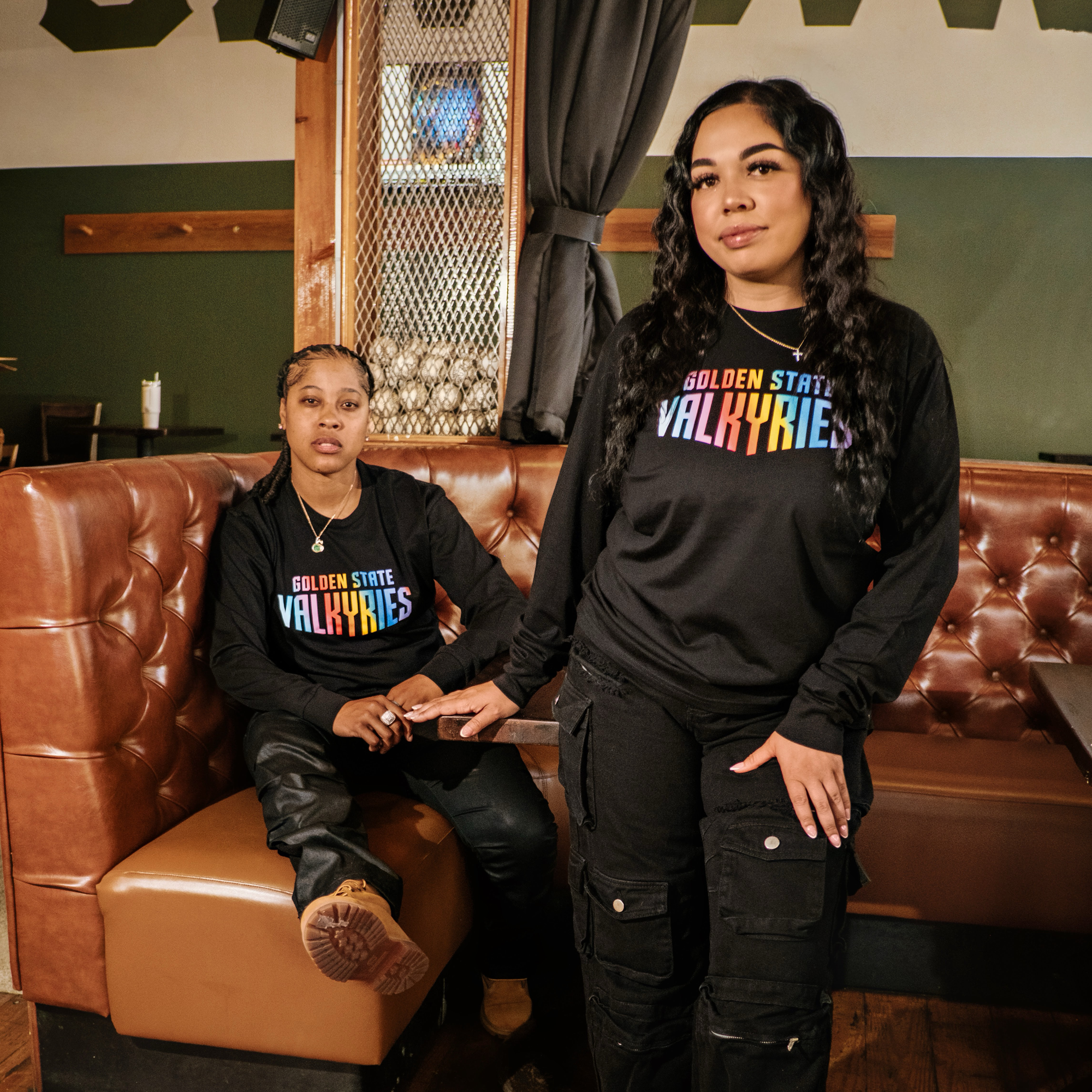 Two women with long dark hair pose hand-in-hand in a cozy, wood-accented cafe, wearing matching black GSV Love LS Tees by GSV. The 100% cotton long sleeve shirts feature rainbow lettering across the front.
