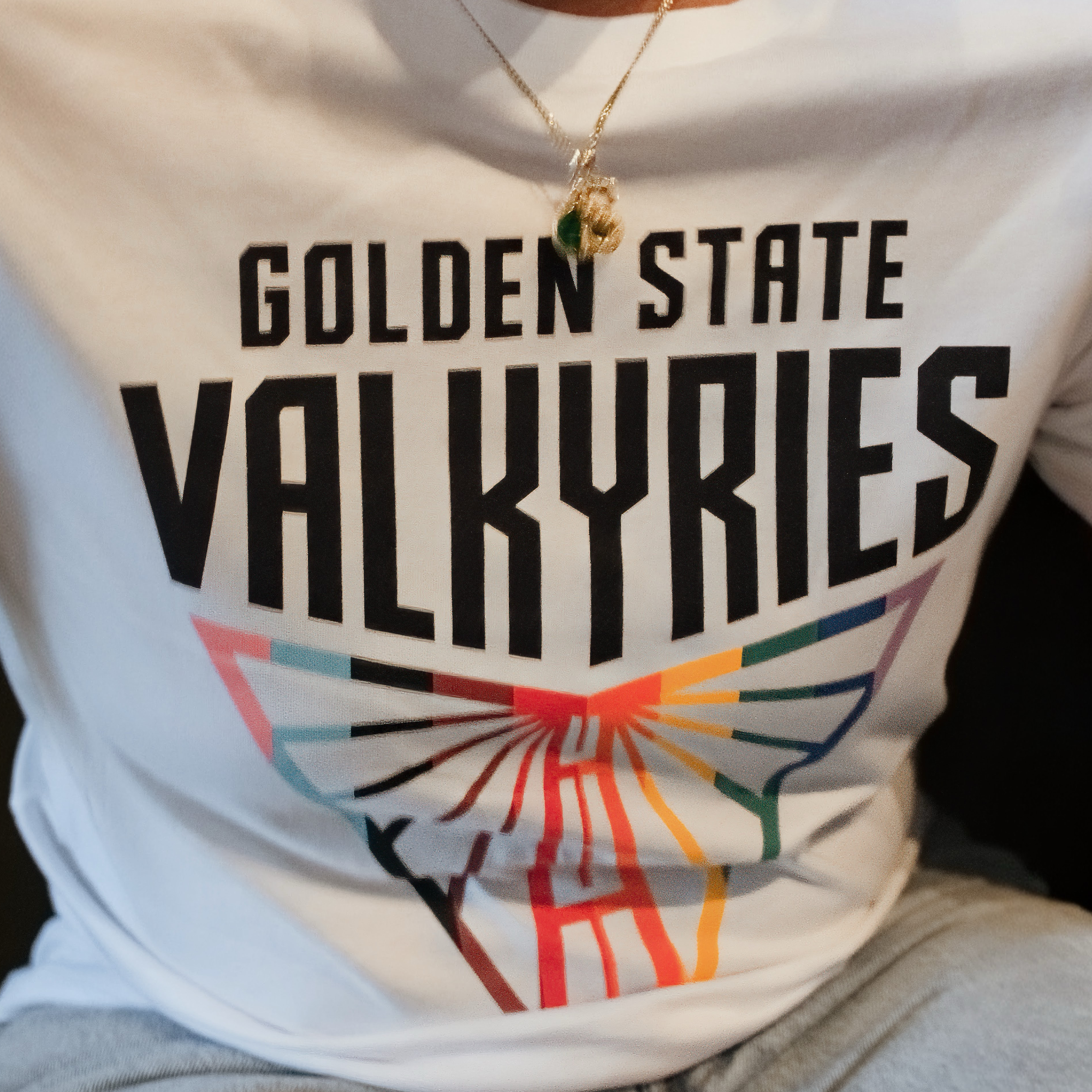 A person wears the GSV Logo Love Tee by GSV, a 100% cotton, classic fit shirt featuring "GOLDEN STATE VALKYRIES" in bold black letters with a vibrant abstract wing design. They also have a gold necklace with a green pendant; only their torso shows.