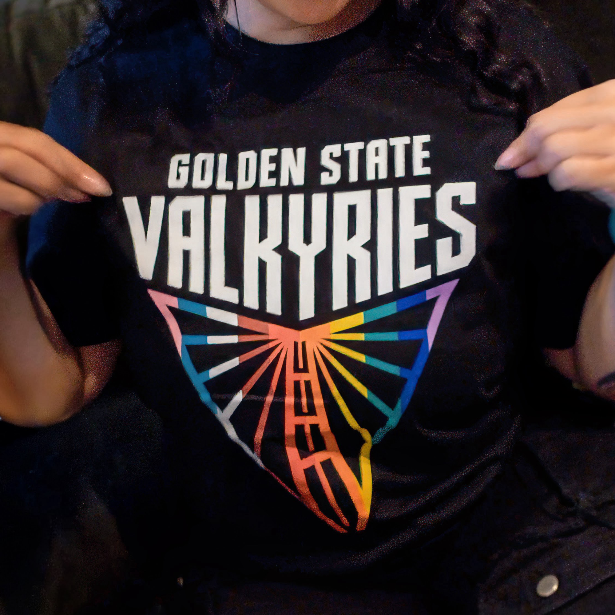A person points to the GSV Logo Love Tee by GSV, a classic fit, 100% cotton t-shirt with bold “Golden State Valkyries” text and a triangular rainbow wing logo. Only their lower face and hands are shown.