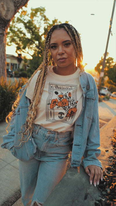 A woman sitting on an Oakland street at sunset leaning against a black car wearing an Oaklandish Blossom t-shirt.