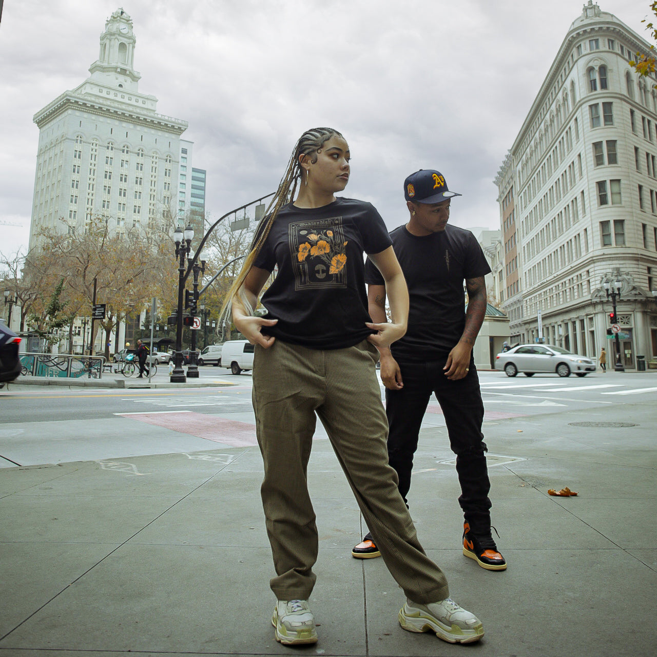 Two individuals stand on a city sidewalk with tall buildings in the background. The person on the left has braided hair, wears an Oaklandish Blossom Tee featuring California poppies, beige pants, and white sneakers. The person on the right dons a black t-shirt, black pants, black hat, and black and orange sneakers.