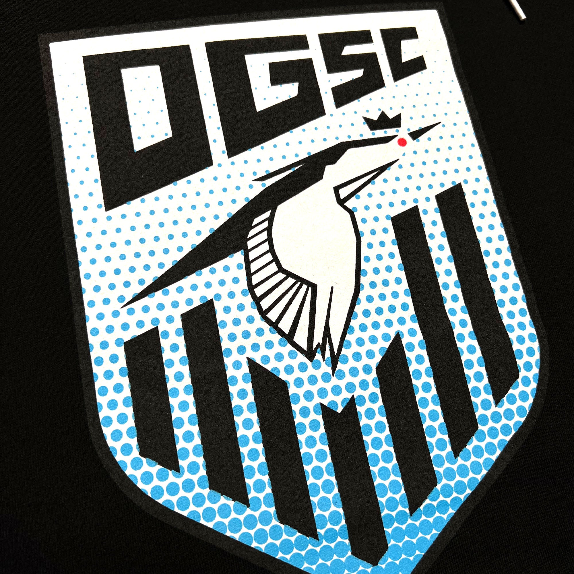 The Oakland Genesis Heron Hoodie by Partner showcases a stylized "OGSC" logo at the top. Beneath this text, a white crowned heron head with a red eye stands out in the center of a blue polka-dot patterned shield. The lower half of the shield features black vertical stripes of varying lengths on a black background, representing Oakland Genesis, a nonprofit organization dedicated to youth soccer.
