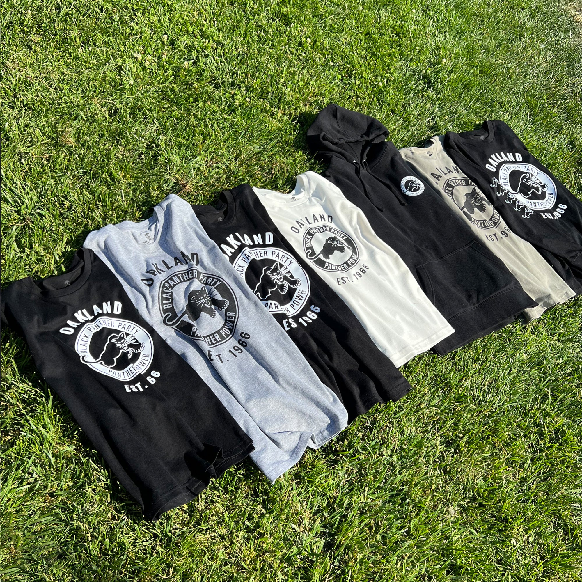 Seven Oaklandish Women's Black Panther Power Tees and one Black Panther Power hoodie are laid out on the grass, each showcasing a black-and-white design with "OAKLAND" and "EST. 1969" framing an image of a hand holding a rose. The tees come in black, gray, and cream, while the predominantly black hoodie radiates Black Panther Power with the same design.