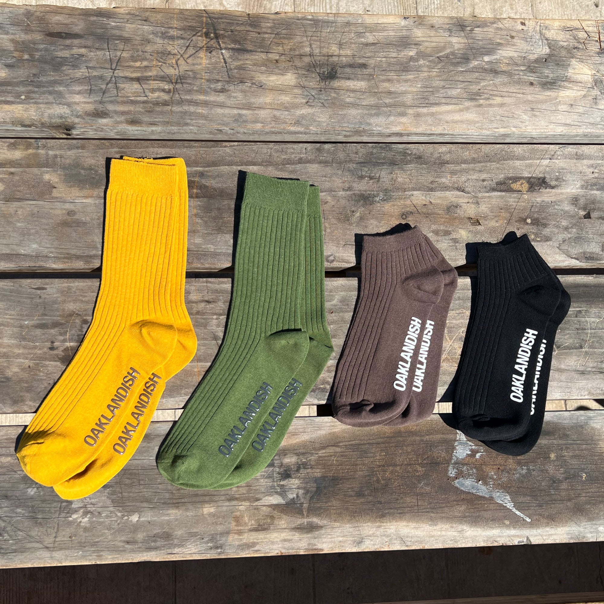 Four pairs of Grip Low Crew Socks are laid out on a rustic wooden surface. The socks, featuring a ribbed design, come in four colors: yellow, olive green, brown, and black. Each pair has the brand name "Oaklandish" printed in bold white letters along the side. The socks appear neatly arranged in a row.
